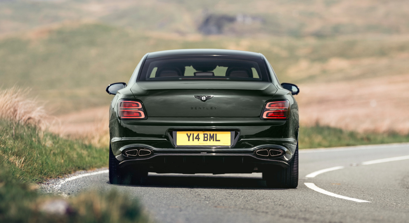 A sleek, dark green luxury sedan with chrome accents on a winding rural road. The car, viewed from the rear, has a license plate reading "Y14 BML."